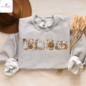 May include: A light gray sweatshirt featuring an embroidered design of four ducks in fall attire. The ducks are depicted with a basket, coffee cup, sweater, and scarf, surrounded by pumpkins and autumn leaves. The sweatshirt has a crew neck.