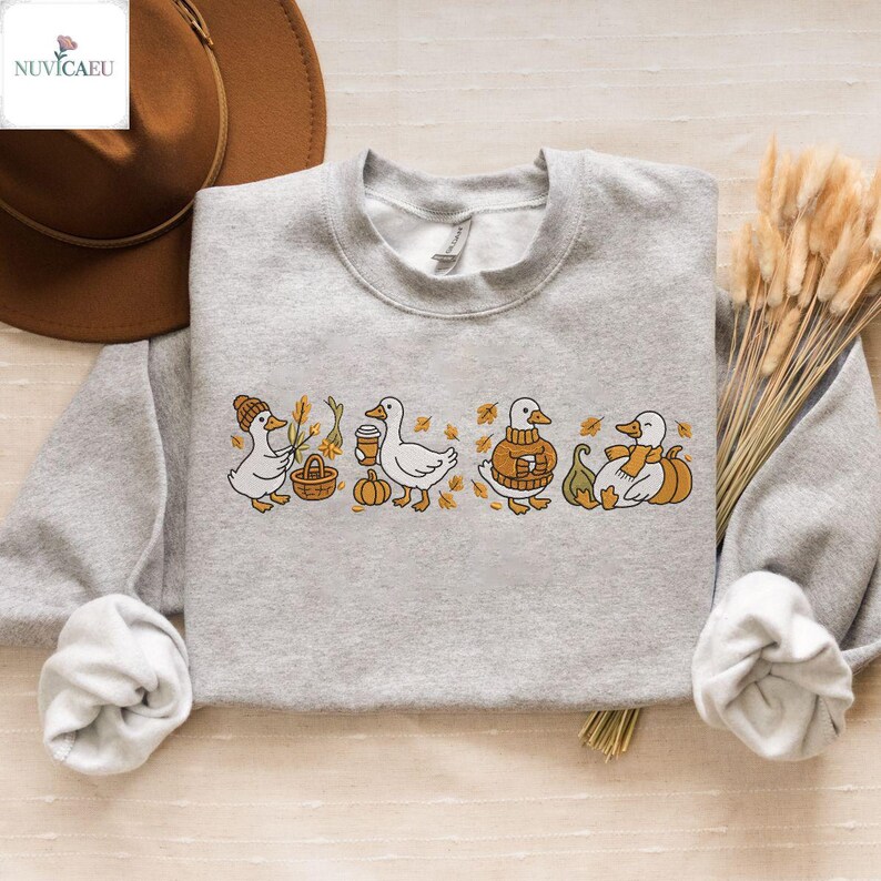 May include: A light gray sweatshirt featuring an embroidered design of four ducks in fall attire. The ducks are depicted with a basket, coffee cup, sweater, and scarf, surrounded by pumpkins and autumn leaves. The sweatshirt has a crew neck.