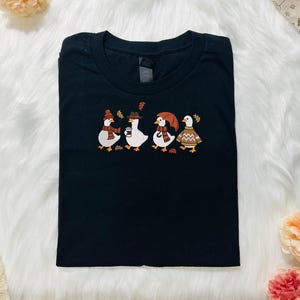 Może przedstawiać: Czarny t-shirt z haftowanym wzorem czterech kaczek w jesiennych strojach. Kaczki mają na sobie czapki, szaliki i swetry, z jesiennymi liśćmi. Koszulka jest złożona na białej, puszystej powierzchni.