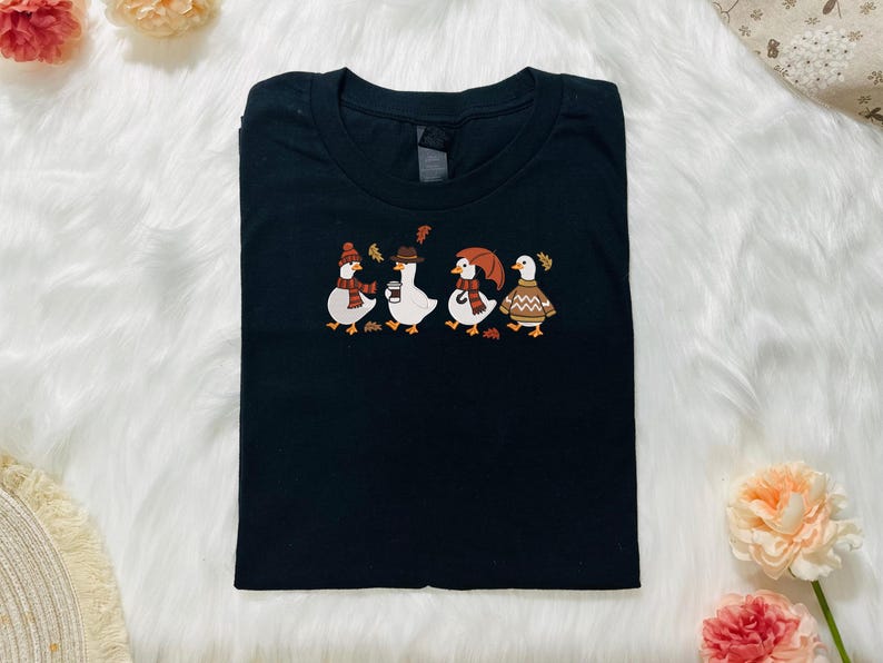 Może przedstawiać: Czarny t-shirt z haftowanym wzorem czterech kaczek w jesiennych strojach. Kaczki mają na sobie czapki, szaliki i swetry, z jesiennymi liśćmi. Koszulka jest złożona na białej, puszystej powierzchni.