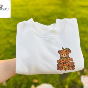 Könnte beinhalten: Ein weißes Sweatshirt mit einem gestickten Design eines Teddybären, der als Halloween-Kürbis verkleidet ist. Der Bär ist braun mit einem Kürbiskopf und sitzt auf Herbstblättern. Das Sweatshirt hat einen Rundhalsausschnitt.