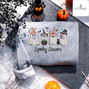 Könnte beinhalten: Ein graues Sweatshirt mit dem gestickten Schriftzug "Spooky Season" und vier Cartoon-Geistern mit Hexenhüten. Eine schwarze Katze und ein Kessel sind ebenfalls auf dem Sweatshirt gestickt. Die Ärmel sind umgeschlagen.