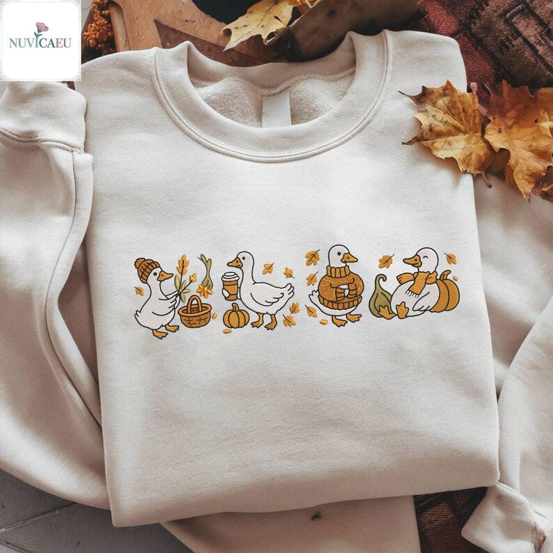 May include: Cream-colored sweatshirt featuring an embroidered design of four ducks in autumnal attire. The ducks are depicted with fall-themed accessories like a hat, coffee cup, and pumpkin. The design includes fall leaves and pumpkins.