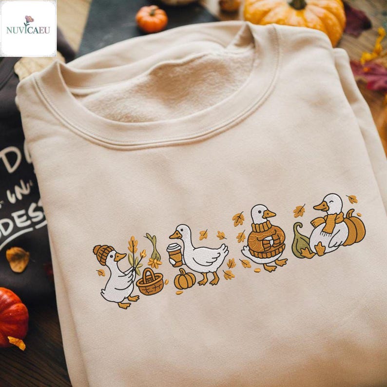May include: Beige sweatshirt featuring an embroidered design of four ducks in autumn attire. One duck wears a hat and carries a basket, another holds a coffee cup, and the others are dressed in a sweater and scarf, with pumpkins and leaves.