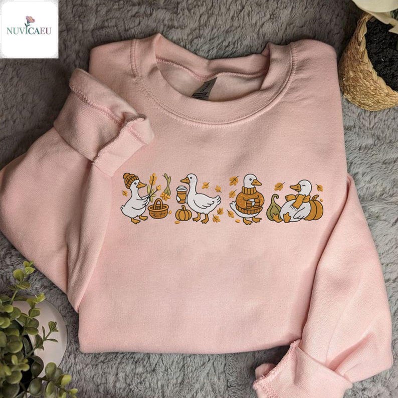 May include: A light pink sweatshirt featuring a line of cartoon ducks in fall attire. One duck wears a hat and carries a basket, another holds a coffee cup, and others are in sweaters and scarves. The design includes pumpkins and fall leaves.