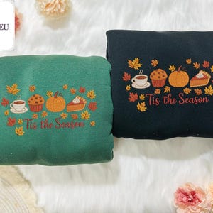 May include: Two sweatshirts, one green and one black, embroidered with fall-themed designs. Each features a coffee cup, muffin, pumpkin, and pie, surrounded by autumn leaves, with the text "Tis the Season" below. The sweatshirts are displayed on a white, fluffy surface.