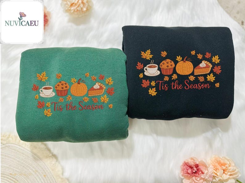 May include: Two sweatshirts, one green and one black, embroidered with fall-themed designs. Each features a coffee cup, muffin, pumpkin, and pie, surrounded by autumn leaves, with the text "Tis the Season" below. The sweatshirts are displayed on a white, fluffy surface.