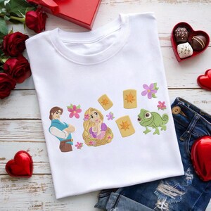 Può includere: T-shirt bianco con personaggi dei cartoni animati ricamati ed elementi di una fiaba. Il design include una principessa con lunghi capelli biondi, un principe, lanterne, fiori e un camaleonte verde. La maglietta è esposta su una superficie di legno con jeans.