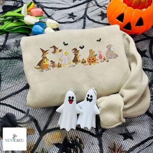 Può includere: Felpa beige a tema Halloween con un design ricamato di personaggi dei cartoni animati in costume. Due portachiavi a forma di fantasma bianco sono davanti alla felpa. Una zucca arancione intagliata e dei fiori sono sullo sfondo.