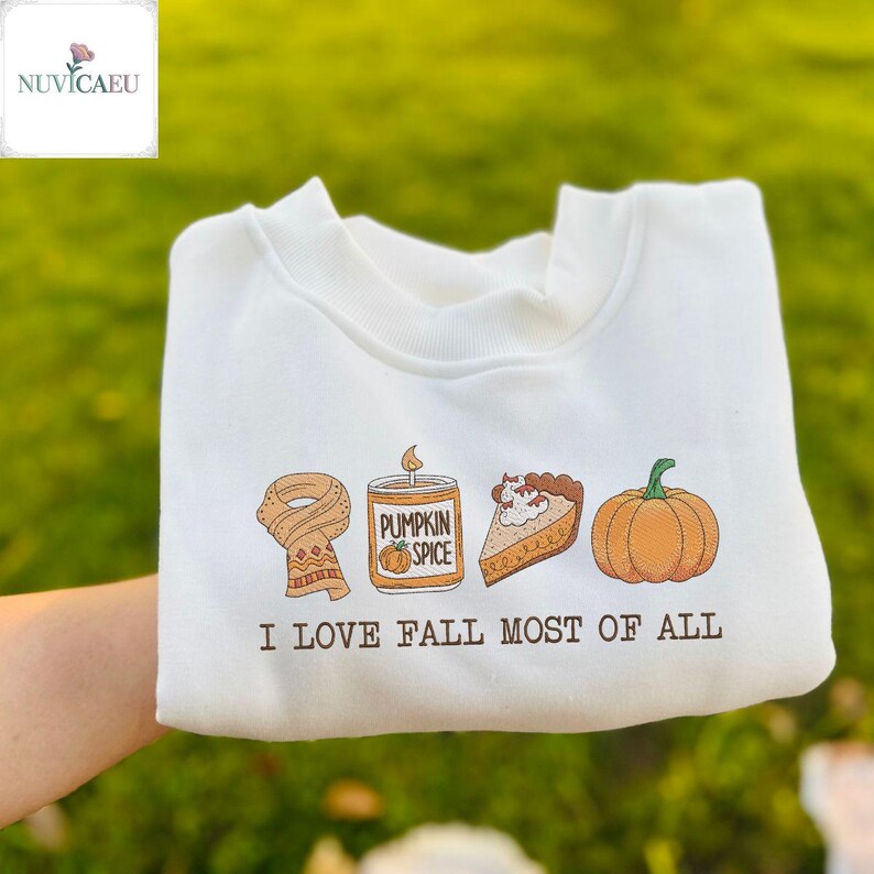 May include: White sweatshirt with an autumn-themed graphic. The design includes a scarf, pumpkin spice candle, pumpkin pie slice, and a pumpkin. The text "I LOVE FALL MOST OF ALL" is printed below the graphic.