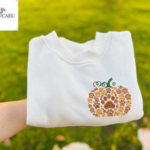 Könnte beinhalten: Weißes Sweatshirt mit einem gestickten Kürbismotiv aus Hundepfotenabdrücken in Braun- und Orangetönen. Der Kürbis hat einen grünen Stiel und Ranken. Das Sweatshirt ist gefaltet, mit dem Markenlogo in der oberen linken Ecke.
