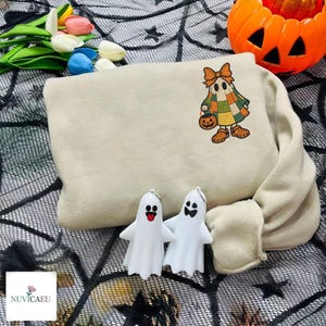 Könnte beinhalten: Beiges Halloween-Sweatshirt mit einem gestickten Geisterdesign, das einen Kürbis hält. Zwei kleine, weiße, geisterförmige Schlüsselanhänger befinden sich vor dem Sweatshirt. Ein orangefarbener Kürbis und Blumen sind im Hintergrund. Das Sweatshirt hat einen langen Ärmel.