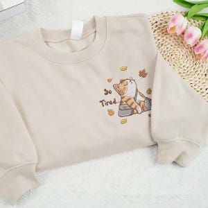 Könnte beinhalten: Beiges Sweatshirt mit gesticktem Design einer schlafenden Katze, umgeben von Herbstblättern. Der Text "So Tired..." ist darunter gestickt. Das Sweatshirt hat einen Rundhalsausschnitt und lange Ärmel.