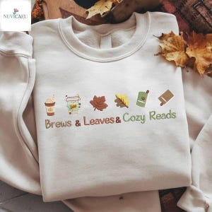 Può includere: Felpa color crema con grafica ricamata a tema autunnale. Presenta una tazza di caffè, un bicchiere da asporto, due foglie, un libro e la scritta "Brews & Leaves & Cozy Reads" in marrone e verde.