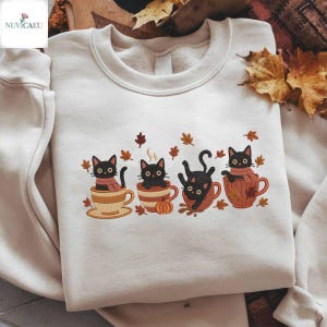 Könnte beinhalten: Cremefarbenes Sweatshirt mit einem gestickten Design von vier schwarzen Katzen in Kaffeetassen und Herbstblättern. Die Katzen sind in verschiedenen Posen, mit Kürbis- und Dampfdetails. Ein gemütliches Herbst-Themen-Design.