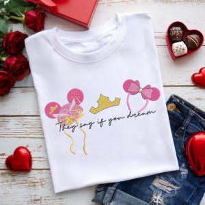 Könnte beinhalten: Weißes T-Shirt mit einem verspielten Design. Es zeigt rosa Mauseohren-Haarbänder, eine goldene Krone und den Text "They say if you dream". Das Design enthält rosa und goldene Akzente, passend für einen lässigen, lustigen Look.