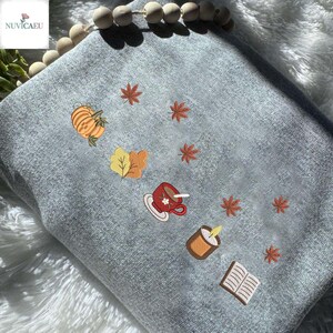 May include: A light gray blanket embroidered with autumn-themed designs. The embroidery includes a pumpkin, fall leaves, a cup of coffee, a candle, and an open book. The blanket is decorated with wooden beads.
