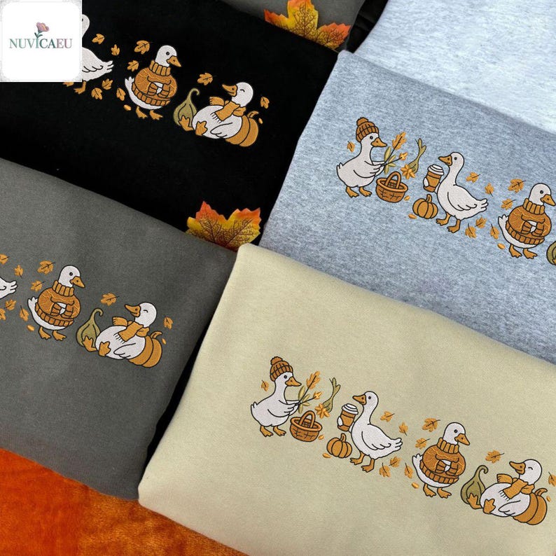 May include: A collection of sweatshirts in various colors, each featuring an embroidered design of ducks in fall attire. The ducks are depicted with pumpkins, leaves, and cozy sweaters, creating a seasonal theme.