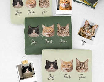 以下が含まれることがあります： 猫の肖像画が描かれた3枚のスウェットシャツ。各スウェットシャツには、3匹の猫の顔と、下に「Jay」、「Tenet」、「Finn」という名前が描かれています。スウェットシャツは、ライトグリーンとダークグリーンです。猫のポラロイド写真が背景にクリップで留められています。