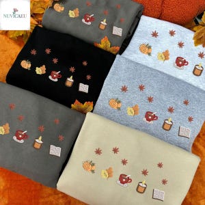 May include: A collection of sweatshirts in various colors, including black, gray, and beige. Each sweatshirt features embroidered autumn-themed designs such as pumpkins, leaves, coffee cups, candles, and books. The sweatshirts are folded and arranged for display.