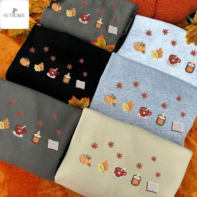 May include: A collection of sweatshirts in various colors, including black, gray, and beige. Each sweatshirt features embroidered autumn-themed designs such as pumpkins, leaves, coffee cups, candles, and books. The sweatshirts are folded and arranged for display.