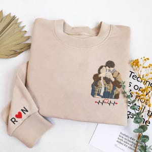 Könnte beinhalten: Beiges Sweatshirt mit einer gestickten Darstellung eines Paares, das ein Selfie macht. Das Design beinhaltet eine Herzschlaglinie und ein rotes Herz. Der linke Ärmel hat die Buchstaben "R ❤️ N" in Schwarz gestickt.