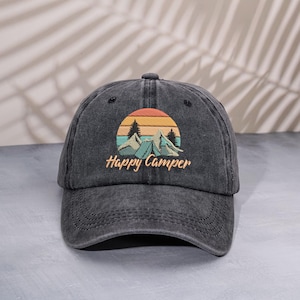 Puede incluir: Gorra de béisbol gris oscuro con visera curva. La gorra presenta un diseño bordado de una puesta de sol sobre montañas y tiendas de campaña, con las palabras "Happy Camper" en letra cursiva.