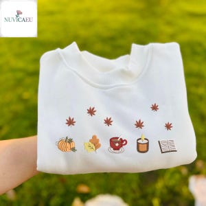May include: White sweatshirt with embroidered autumn-themed designs. Features a pumpkin, leaves, a cup of coffee, a candle, and a book. The design includes small red maple leaves scattered across the top. The brand name "NUVICAEU" is in the top left corner.
