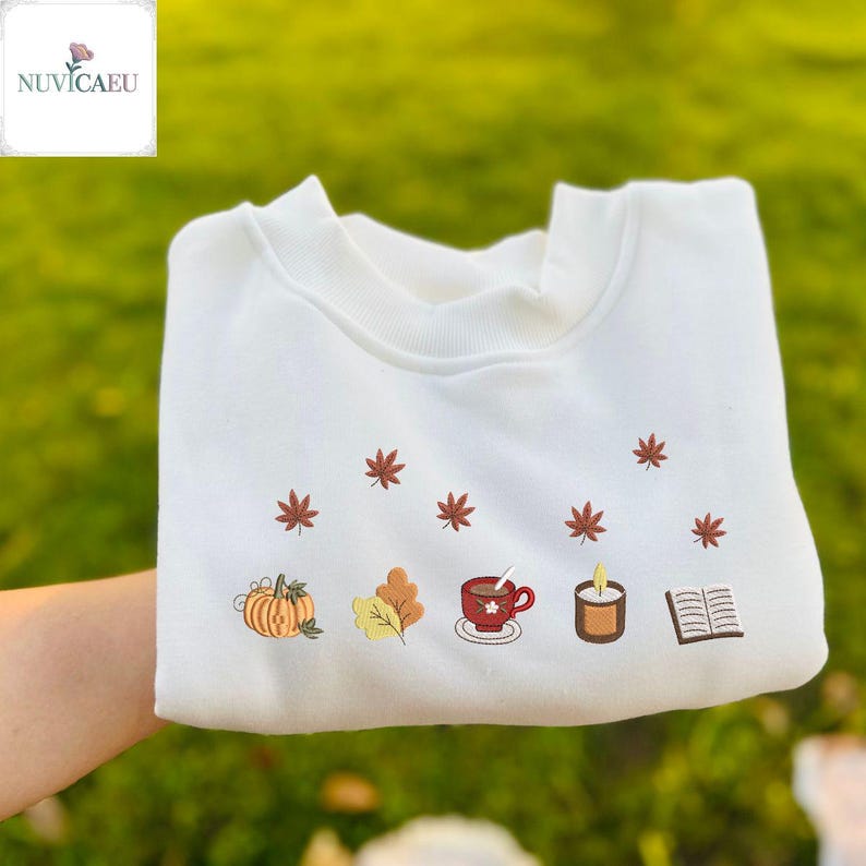 May include: White sweatshirt with embroidered autumn-themed designs. Features a pumpkin, leaves, a cup of coffee, a candle, and a book. The design includes small red maple leaves scattered across the top. The brand name "NUVICAEU" is in the top left corner.