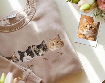 May include: Beige sweatshirt featuring embroidered cat faces with names "Doe", "Oli", and "Jay". A polaroid photo of a ginger cat is next to the sweatshirt. The sweatshirt has a crew neck and long sleeves. A second sweatshirt sleeve is visible.