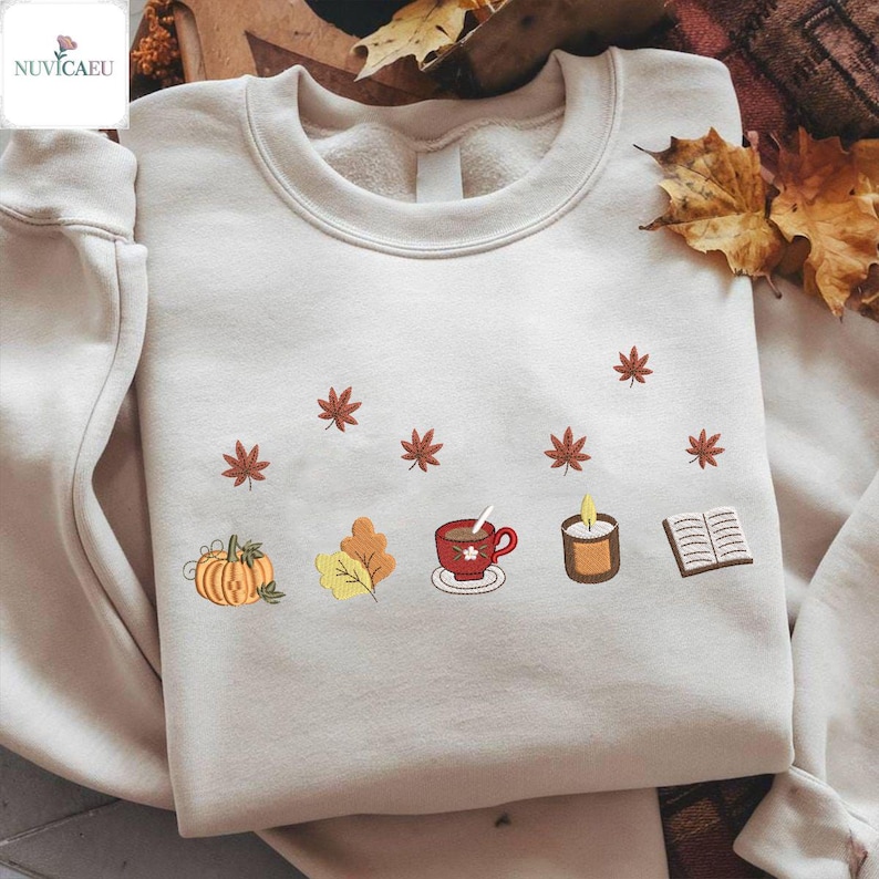 May include: Cream-colored sweatshirt with embroidered autumn-themed designs. Features a pumpkin, leaves, a cup of tea, a candle, and a book. Brown maple leaves are scattered around the design. A cozy fall-themed apparel item.