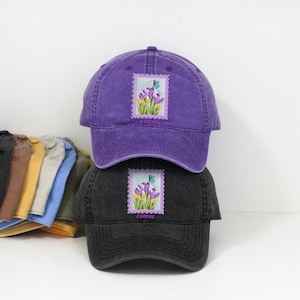 Könnte beinhalten: Zwei Baseballkappen, eine lila und eine schwarze, sind gestapelt. Jede Kappe hat ein gesticktes Stempeldesign mit lila und gelben Blumen, einem blauen Schmetterling und dem Wort "CROCUS". Die Kappen sind aus einem weichen, strukturierten Stoff.