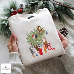 Könnte beinhalten: Cremefarbenes Sweatshirt mit einem gestickten Weihnachtsbaum-Design mit verschiedenen Katzen. Der Baum ist mit Ornamenten geschmückt, und das Design enthält ein rotes Band und eine Zuckerstange. Das Sweatshirt ist gefaltet.