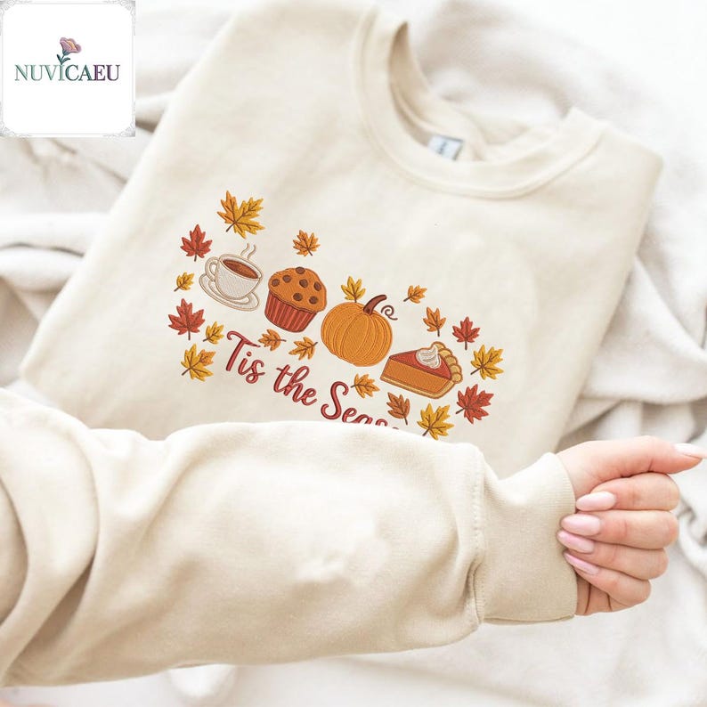 May include: Cream-colored sweatshirt with an embroidered fall-themed design. The design includes a coffee cup, muffin, pumpkin, and pie, surrounded by autumn leaves. The text "Tis the Season" is also embroidered on the sweatshirt.