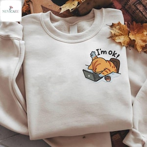 Może przedstawiać: Kremowa bluza z haftowanym wzorem kota pracującego na laptopie. Kot jest pomarańczowo-biały, z napisem "I'm ok!" nad nim. Na biurku stoi filiżanka kawy.