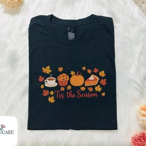 May include: Black t-shirt with an embroidered fall-themed design. The design includes a cup of coffee, a muffin, a pumpkin, and a slice of pie, surrounded by autumn leaves. The text "Tis the Season" is below the design.