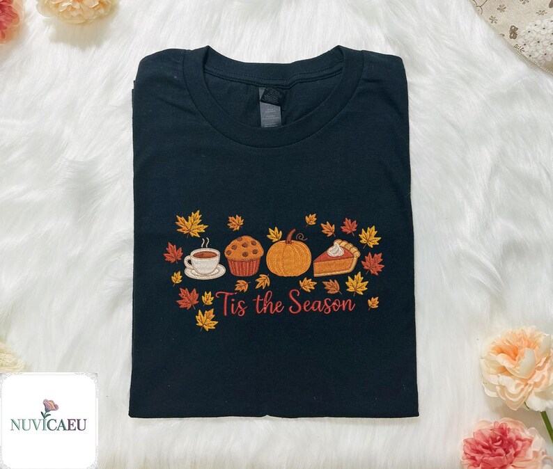 May include: Black t-shirt with an embroidered fall-themed design. The design includes a cup of coffee, a muffin, a pumpkin, and a slice of pie, surrounded by autumn leaves. The text "Tis the Season" is below the design.
