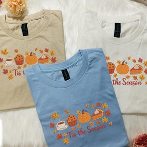 May include: Three t-shirts in beige, light blue, and white, each with a crew neck. The shirts feature a fall-themed graphic with a pumpkin, pie, muffin, coffee cup, and autumn leaves. The text "Tis the Season" is printed on the shirts.