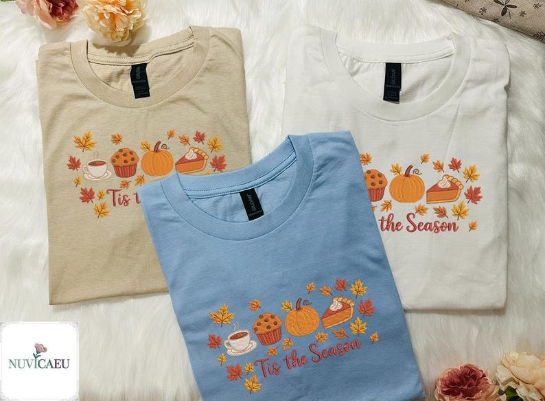 May include: Three t-shirts in beige, light blue, and white, each with a crew neck. The shirts feature a fall-themed graphic with a pumpkin, pie, muffin, coffee cup, and autumn leaves. The text "Tis the Season" is printed on the shirts.