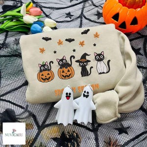 May include: A beige Halloween sweatshirt featuring black cats on pumpkins, bats, and leaves. Two white ghost ornaments and a small pumpkin are also visible. The text "Spooky Season" is embroidered on the sweatshirt.