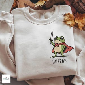 Könnte beinhalten: Cremefarbenes Sweatshirt mit einer gestickten Cartoon-Frosch-Figur, die einen roten Umhang trägt und ein Schwert hält. Das Wort "Huzzah" ist darunter gestickt. Das Sweatshirt hat einen Rundhalsausschnitt und lange Ärmel.