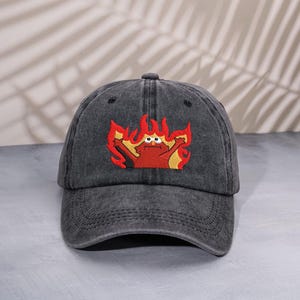 Op de afbeelding: Een donkergrijze baseballpet met een gebogen klep. De pet heeft een geborduurd ontwerp van een rood personage omringd door gele en oranje vlammen. Het ontwerp staat op de voorkant.
