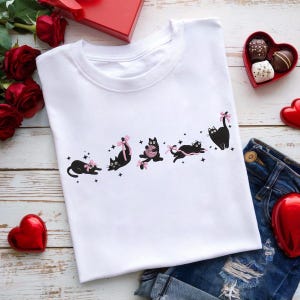 Könnte beinhalten: Weißes T-Shirt mit einem Design aus schwarzen Katzen mit rosa Schleifen und Garn. Die Katzen sind in verschiedenen Posen, mit Sternen verstreut. Das Shirt wird mit Jeansshorts, roten Rosen und herzförmigen Pralinen kombiniert.
