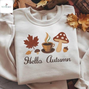 Peut inclure: Sweat-shirt crème avec un motif automnal brodé. Le motif comprend une feuille d'érable marron, une tasse de café, un champignon et le texte "Hello Autumn" en script marron. Un gland et une petite feuille sont également inclus.