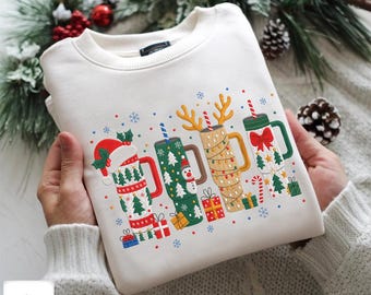 以下が含まれることがあります： クリスマスデコレーションが施されたお祝いのマグカップの刺繍デザインが施されたクリーム色のスウェットシャツ。マグカップには、サンタの帽子、クリスマスツリー、雪だるま、プレゼントが含まれています。デザインは赤、緑、金です。