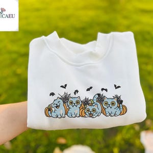 Pode incluir: Camisola branca com um design de Halloween. O design inclui gatos de desenho animado, abóboras, teias de aranha e morcegos. A camisola é segurada por uma mão. A marca "NUVICAEU" está no canto superior esquerdo.
