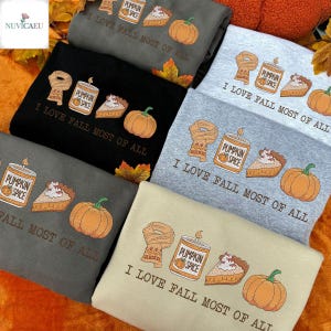 May include: A collection of folded sweatshirts in various colors, each featuring a fall-themed design. The design includes a scarf, a pumpkin spice candle, a slice of pie, and a pumpkin, with the text "I LOVE FALL MOST OF ALL".