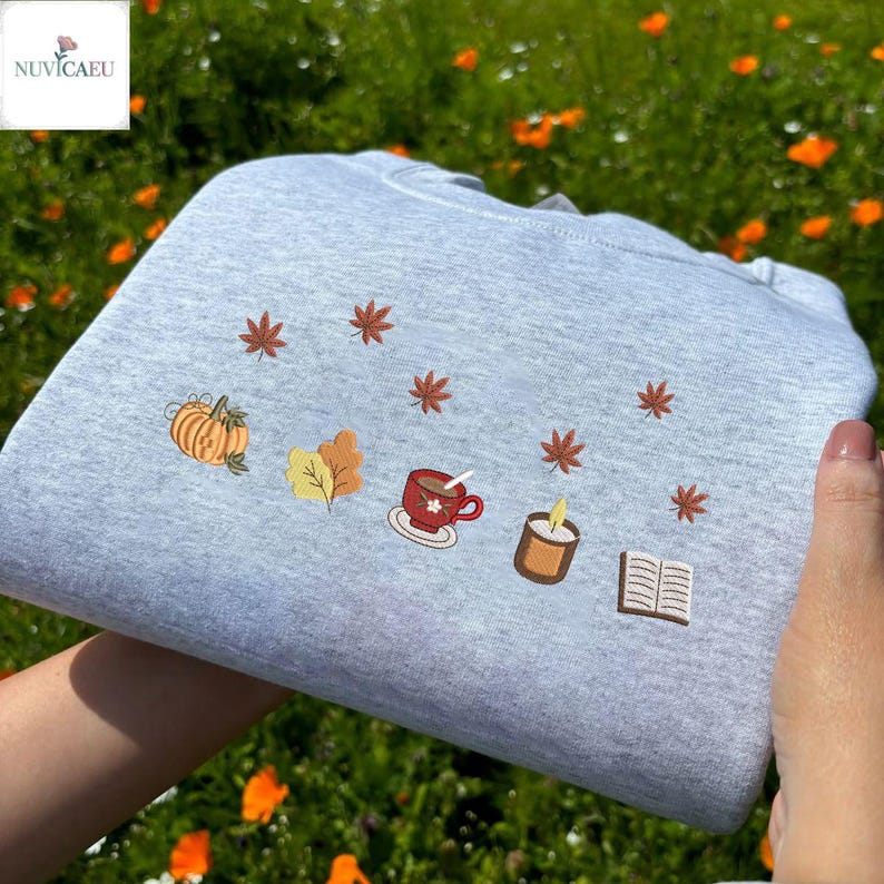 May include: A light gray sweatshirt with embroidered autumn-themed designs. The design includes a pumpkin, leaves, a cup of coffee, a candle, and a book, with several small leaves scattered around the design.