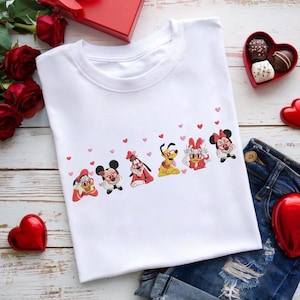 Può includere: T-shirt bianco con una fila di personaggi dei cartoni animati, tra cui Topolino e Minnie, e cuori rosa. La maglietta è mostrata con decorazioni di San Valentino, tra cui rose rosse e cioccolatini a forma di cuore.