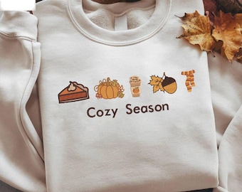 May include: A cream-coloured sweatshirt featuring embroidered autumn-themed motifs: a pie slice, pumpkin, coffee, leaf, acorn, and scarf. The words "Cozy Season" are embroidered below. The sweatshirt is folded on a textured surface.